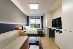 A hotel room with a bed, a desk with a television, a sofa, and a window with a view of greenery. - Sokcho Chonpines Beach Hotel (Sokcho)
