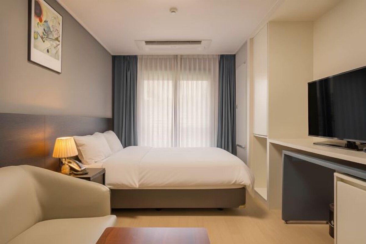 A hotel room with a bed, a desk with a TV, a lamp, and a painting on the wall.