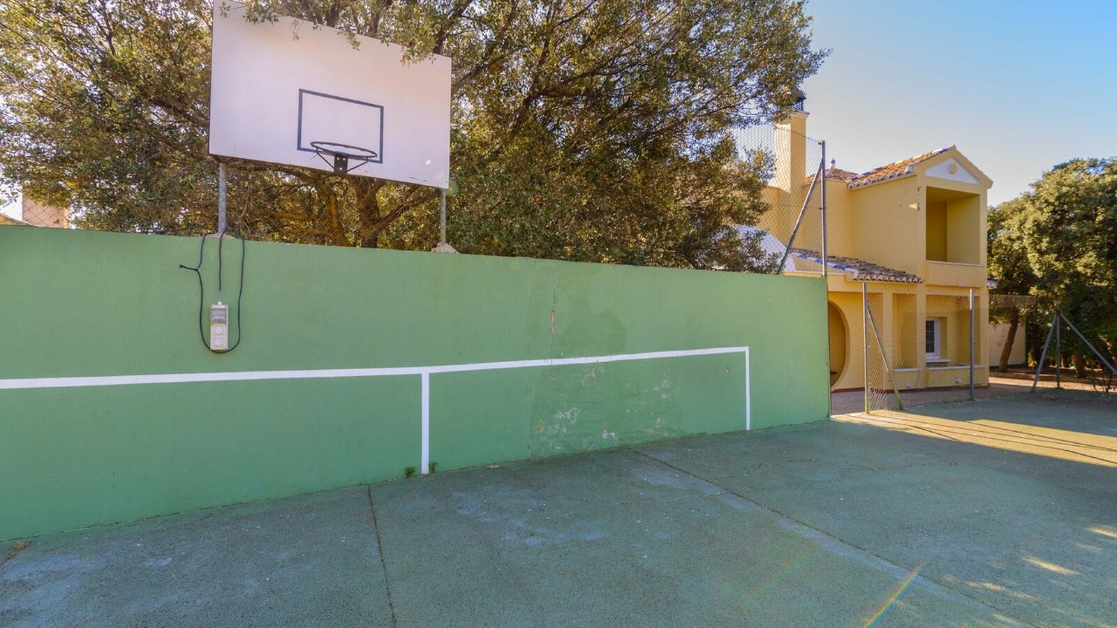 A basketball court with a green wall, a basketball hoop, and a yellow building in the background.