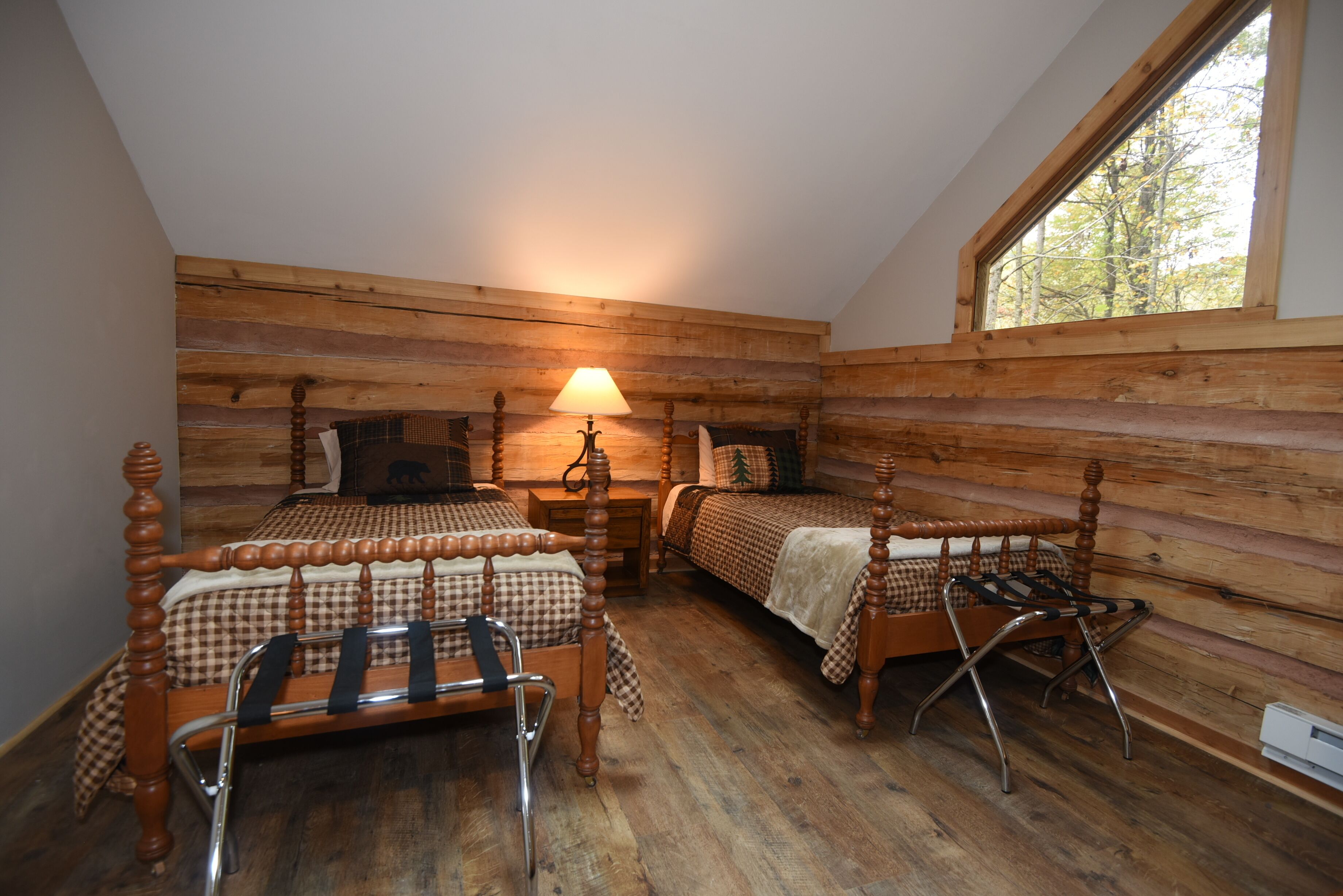 Une chambre dans un chalet en rondins, avec deux lits en bois, une table de chevet, une lampe et une fenêtre donnant sur la forêt.