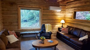 Un interior acogedor de cabaña de madera con un sofá de cuero, una mesa de centro de madera con un jarrón y libros, un sillón de cuero y un ventanal con vistas al bosque.