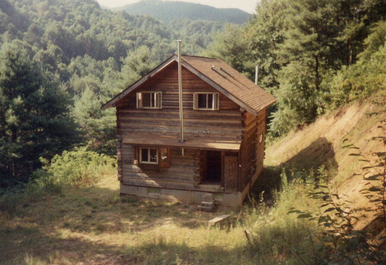 A rustic log cabin with a steeply pitched roof, a front porch, and a chimney.