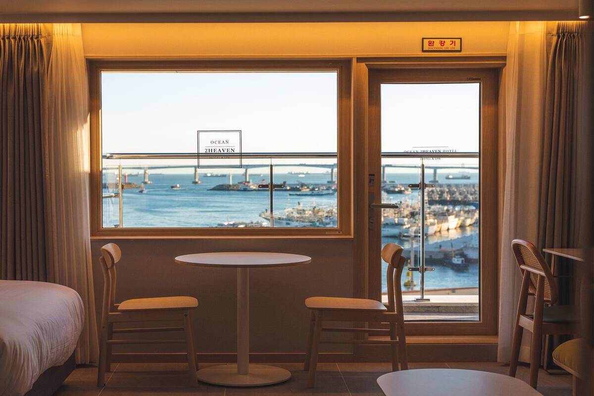A hotel room with a large window offering a view of a bridge and marina. There is a bed, two chairs, and a small table.