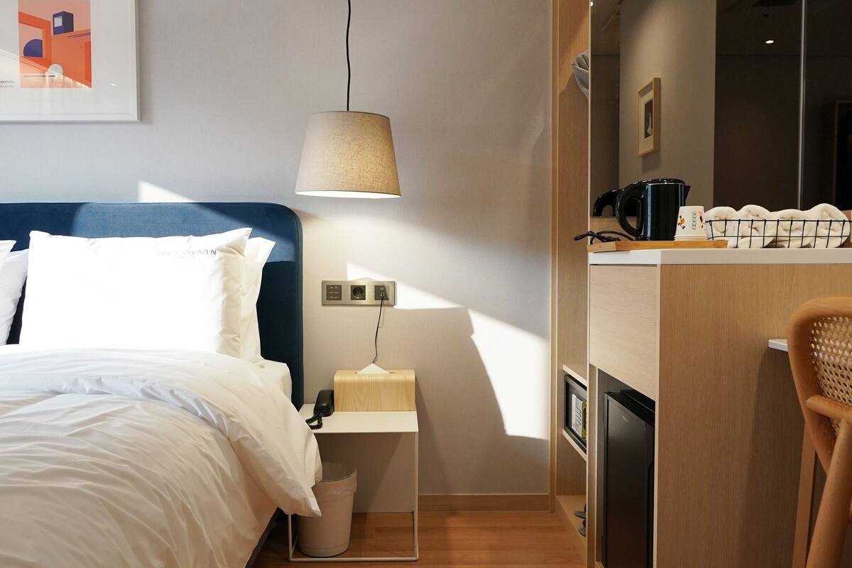 A hotel room with a bed, bedside table, lamp, and a kitchenette area.