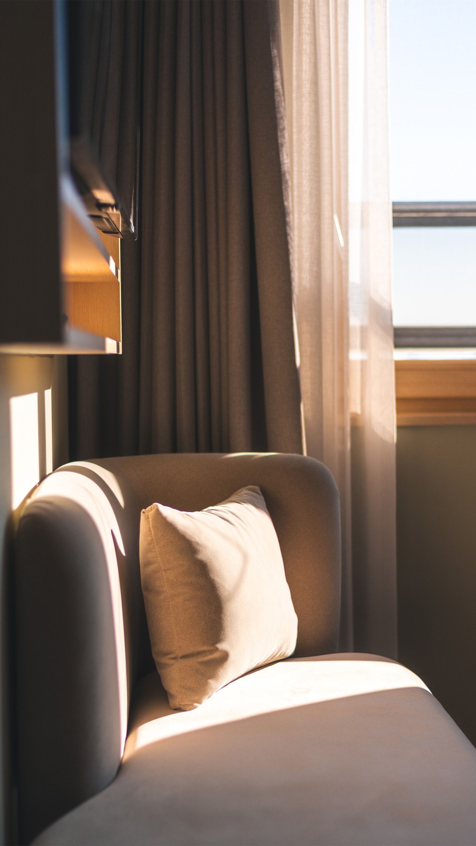 A corner of a room with a window, curtains, and a chair with a pillow.