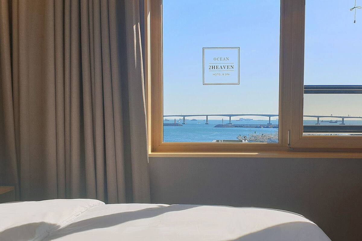 A hotel room with a bed, a window with a view of a bridge and the sea, and a sign reading "Ocean 2Heaven Hotel & Spa".