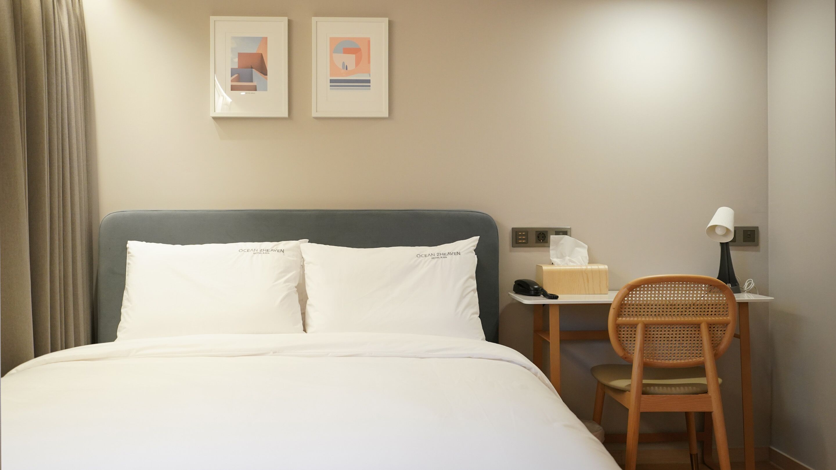 A hotel room with a bed, a desk with a chair, a lamp, a phone, and two framed pictures on the wall.