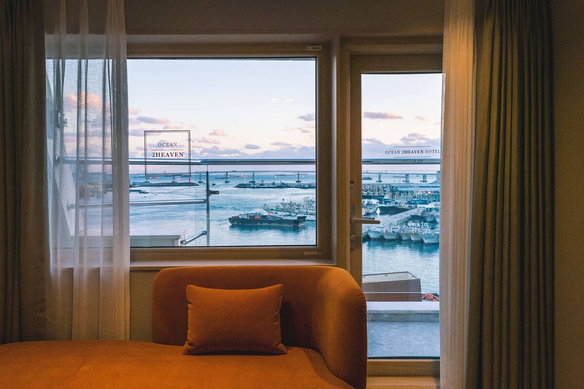 A hotel room with a bed, a window with a view of a harbor, and curtains.