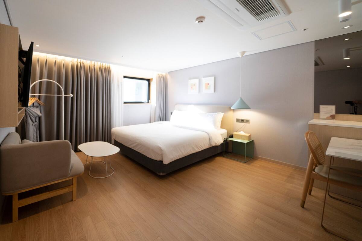 A modern hotel room with a bed, a desk, a chair, a small table, and a wardrobe.