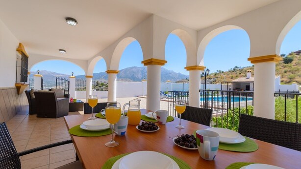 Uma mesa de pequeno-almoço num terraço com vista para a piscina, com pratos, copos e um jarro de sumo de laranja.