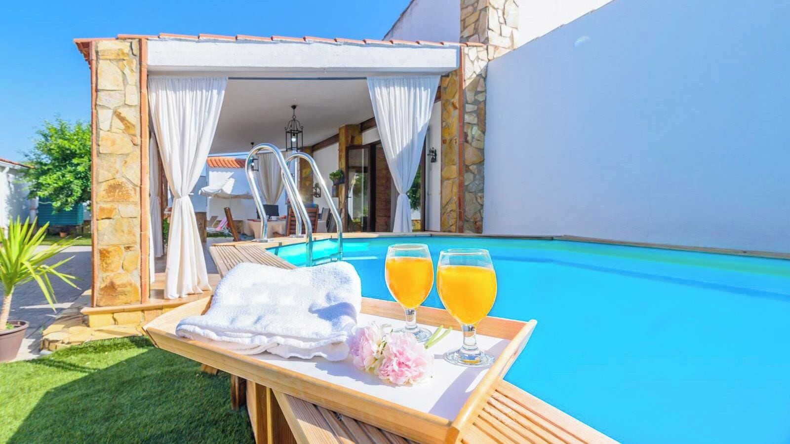 A poolside setting with a wooden table, a towel, two glasses of orange juice, and a flower arrangement.