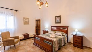Ein Schlafzimmer mit einem hölzernen Bett, zwei Nachttischen mit Lampen, einem Sessel und einem Fenster mit durchsichtigen Vorhängen.