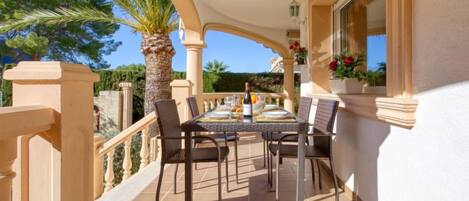 A covered outdoor dining area with a table set for two, surrounded by chairs, palm trees, and a view of the landscape.