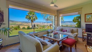 A spacious living room with a large window offering a scenic view of a coastal landscape, a ceiling fan, and a television.