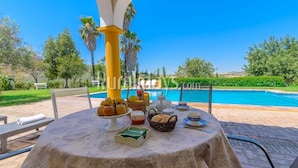 A table set for breakfast with fruits, bread, and tea, overlooking a pool and garden.