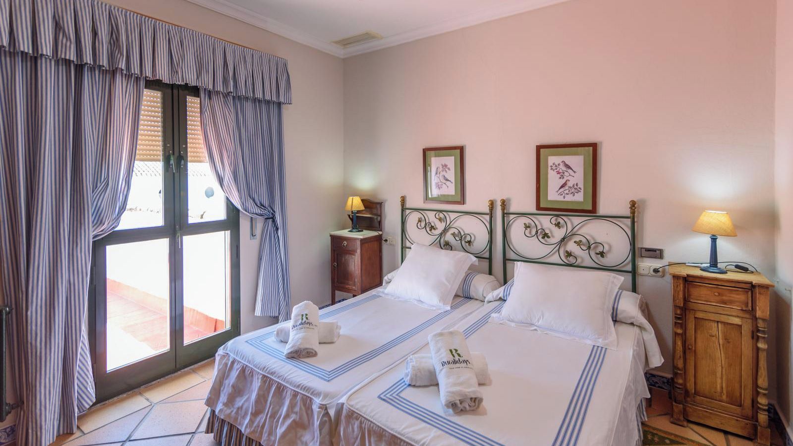 A hotel room with two beds, a window with curtains, a bedside table with a lamp, and a wooden cabinet.