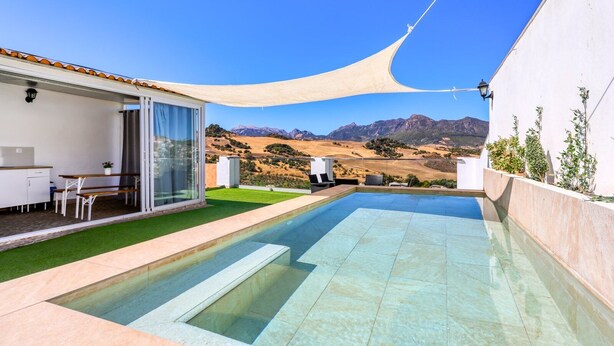 A modern outdoor pool area with a clear view of mountains in the background, a covered seating area, and a glass-enclosed space with a table and chairs. - Casa El Mirador Montecorto by Ruralidays (Málaga)