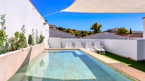A swimming pool with a clear blue water, surrounded by white walls and a white canopy. There are white lounge chairs around the pool.