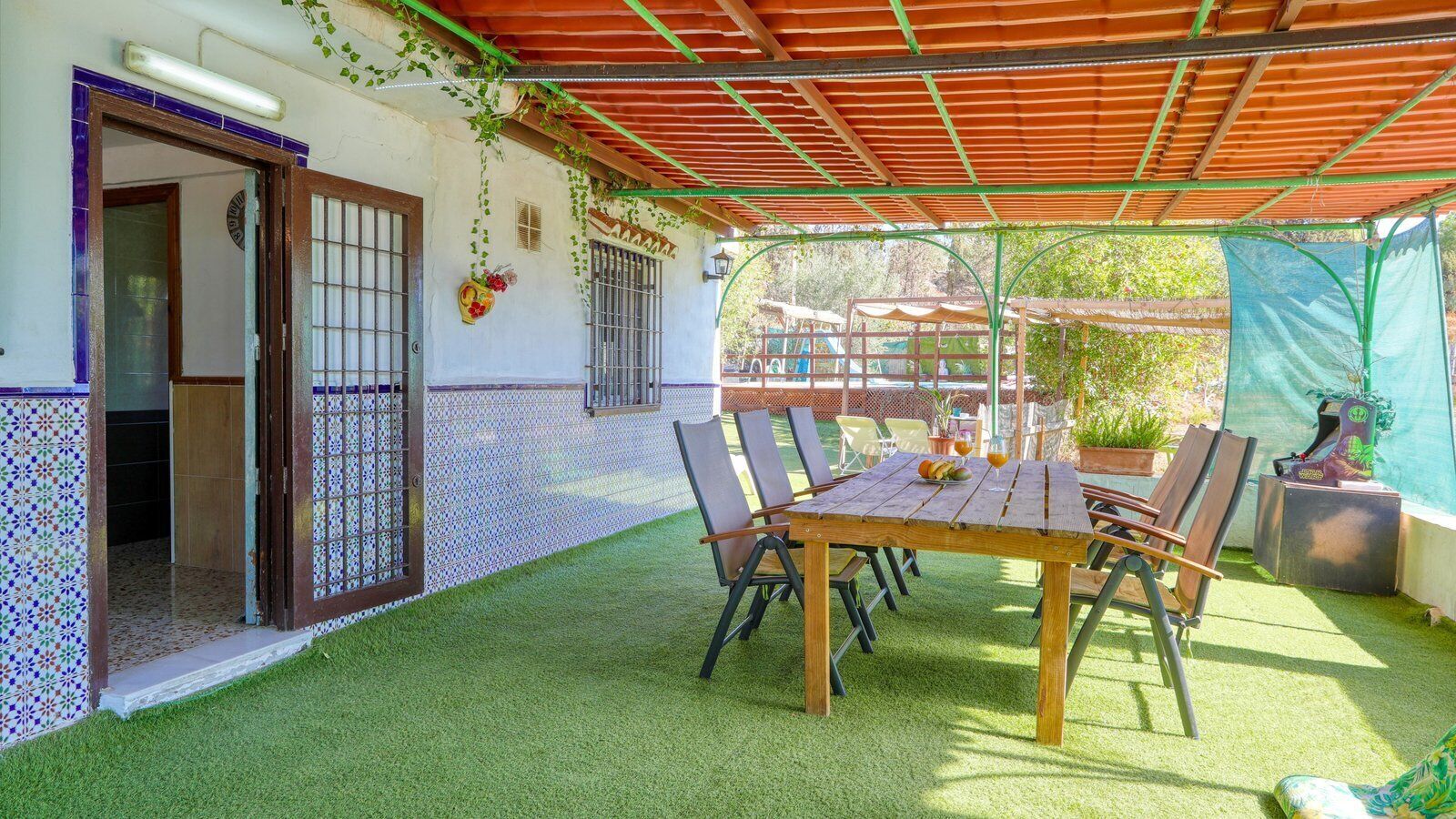 Eine Terrasse mit Holztisch und -stühlen, umgeben von Kunstgrün, einer Pergola und einem eingezäunten Garten.