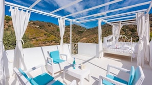 Un espace extérieur aménagé avec une zone salon couverte, doté de meubles en osier blanc et de coussins bleus, d’un lit blanc et offrant une vue sur un paysage vallonné.