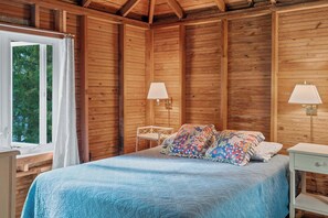 A wooden cabin bedroom with a bed, two bedside lamps, a window with curtains, and a view of trees. - Waterfront, Dog Friendly Sherwood Point (Sturgeon Bay)