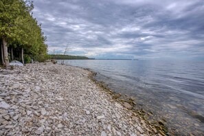 A pebbled beach with trees and a calm lake under a cloudy sky. - Waterfront, Dog Friendly Sherwood Point (Sturgeon Bay)