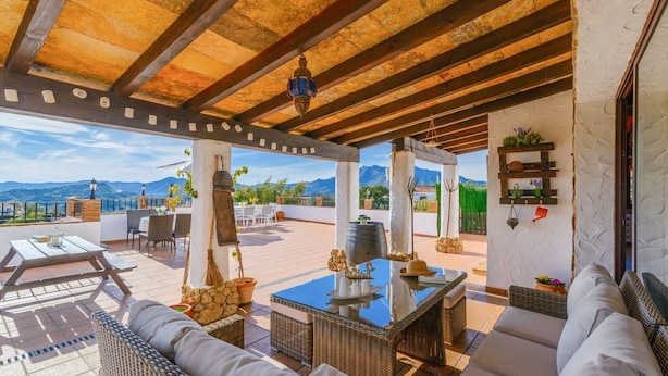 Une terrasse extérieure couverte, aménagée avec des meubles en osier, une table dont le plateau est bleu, et offrant une vue sur des montagnes en arrière-plan.