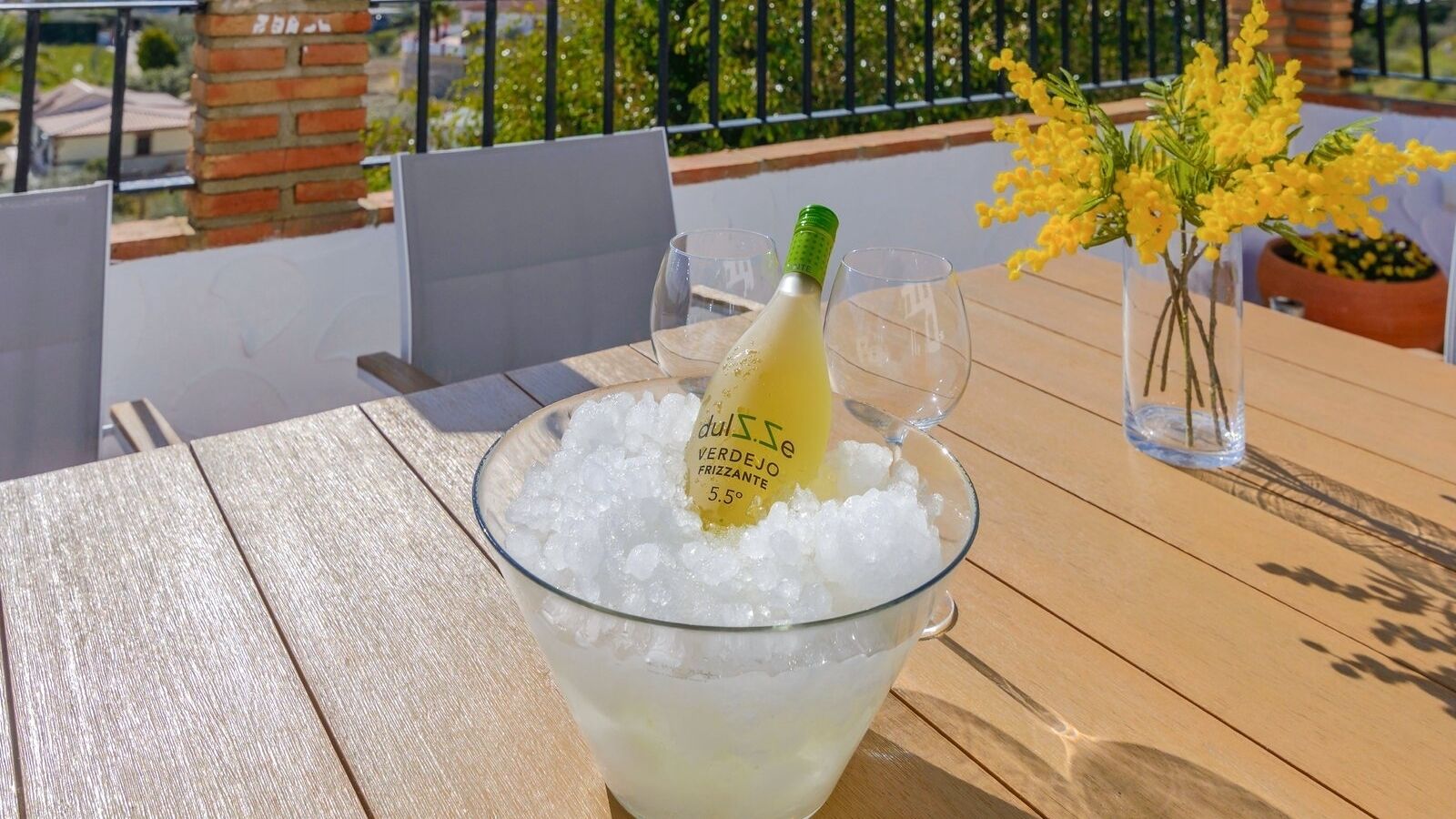 Un tazón de vidrio lleno de hielo, una botella de vino 'duse Verdejo Frizzante 5.5º' y un florero con flores amarillas sobre una mesa de madera.