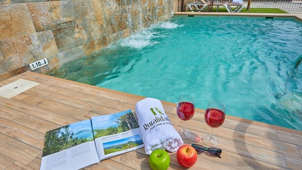 Une terrasse en bois au bord d’une piscine, avec une serviette, deux verres de vin rouge, une pomme et un livre.