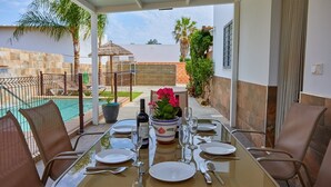 Un espace extérieur aménagé pour les repas, avec une table dressée pour quatre personnes, entourée de chaises, et une piscine ainsi qu’une végétation luxuriante en arrière-plan.