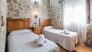 A room with two beds, stone walls, wooden headboards, a nightstand, and a window with sheer curtains.