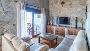 A stone-walled living room with a glass coffee table, beige sofas, a flat-screen TV, and a stone fireplace.