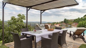 An outdoor patio with wicker furniture, a table set with a basket and glasses, and a view of a hillside with houses.