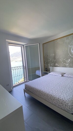 A bedroom with a large bed, a nightstand, and a view of the sea through a sliding glass door.