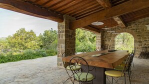 Un patio al aire libre con una mesa de madera larga y sillas, paredes de piedra y vistas al verdor.