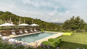 Zona de piscina con sillas de descanso y sombrillas, rodeada de vegetación y colinas.