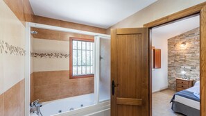 A bathroom with a bathtub, a window, and a wooden door leading to a bedroom with a bed and a nightstand. - Villa Virginia Arenas by Ruralidays (Málaga)