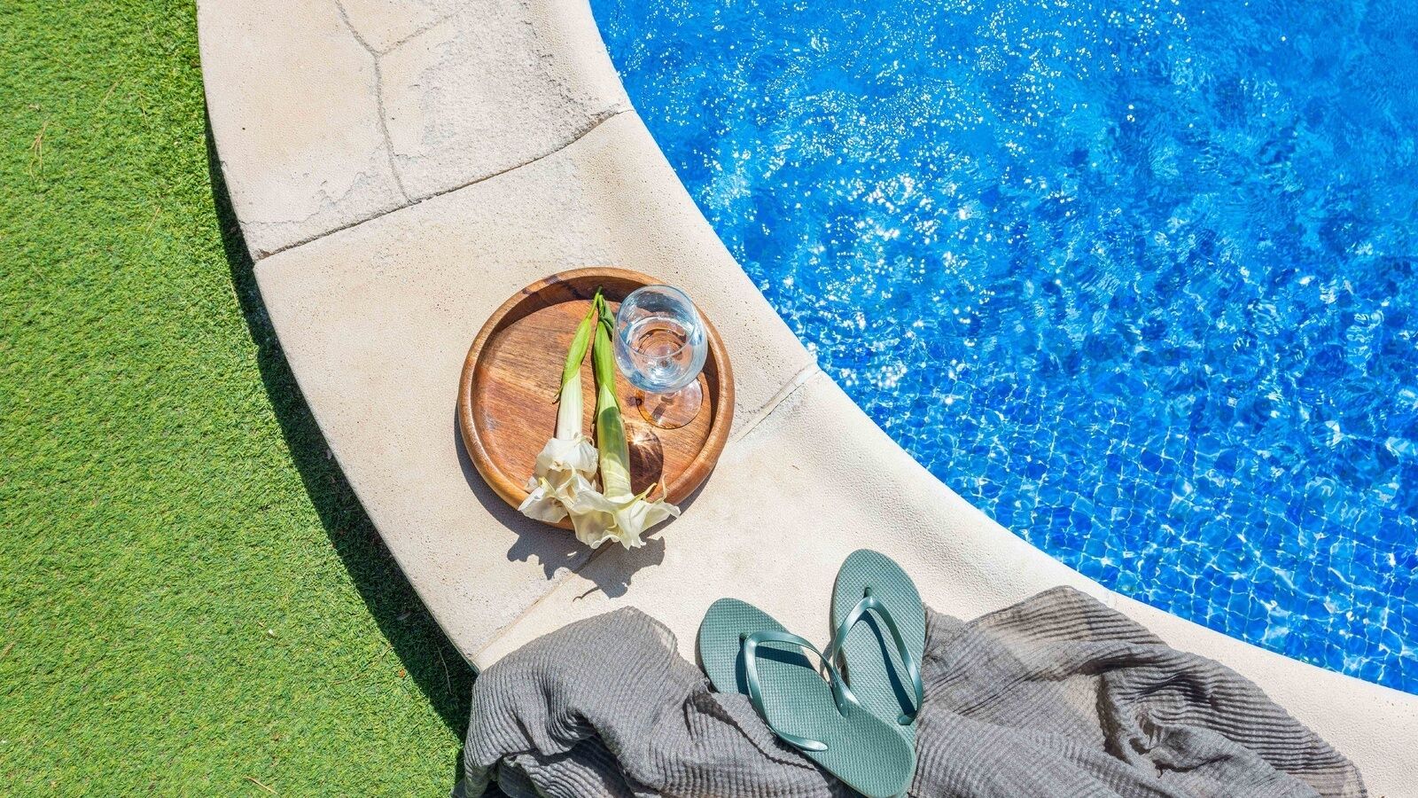 Ett område vid poolen med en kant av betong, ett träbricka med ett glas vatten och grönsaker samt ett par gröna flip-flops.