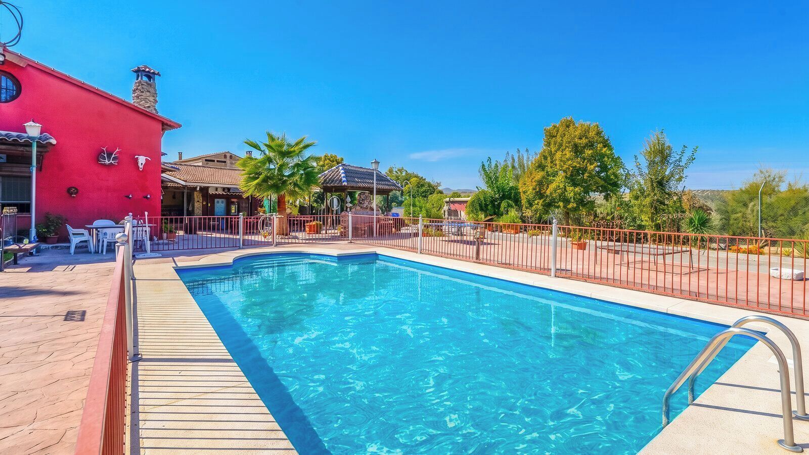 A swimming pool with a wooden deck, surrounded by a fence, and a red building with a chimney in the background.