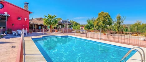 A swimming pool with a wooden deck, surrounded by a fence, and a red building with a chimney in the background.