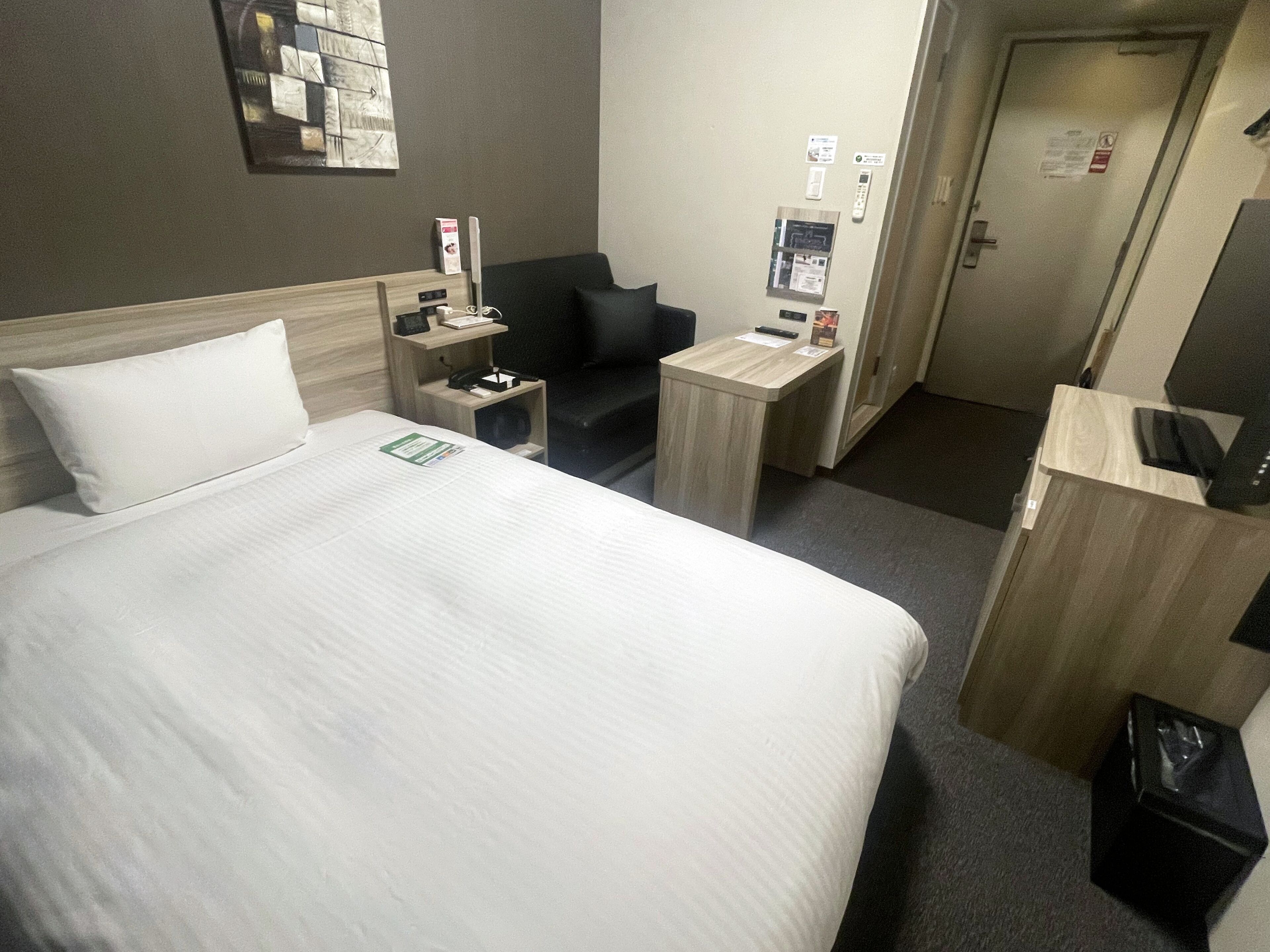 Una habitación de hotel con cama, escritorio, silla, mesita, cubo de basura y puerta.
