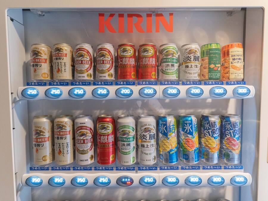 Máquina automática de bebidas com diversas opções, incluindo cerveja Kirin e outras bebidas, com etiquetas de preço e botões de seleção.