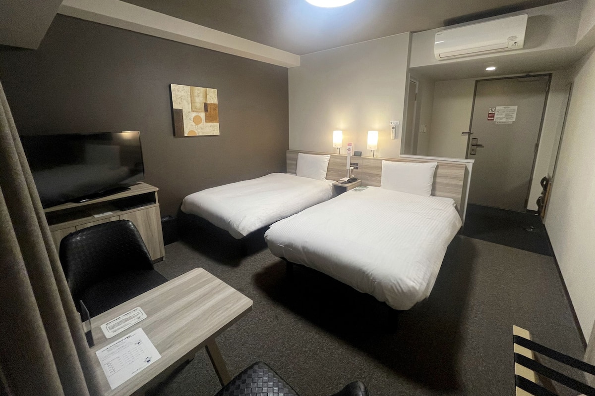 A hotel room with two single beds, a desk with a chair, a flat-screen TV, a wall-mounted artwork, and an air conditioning unit.