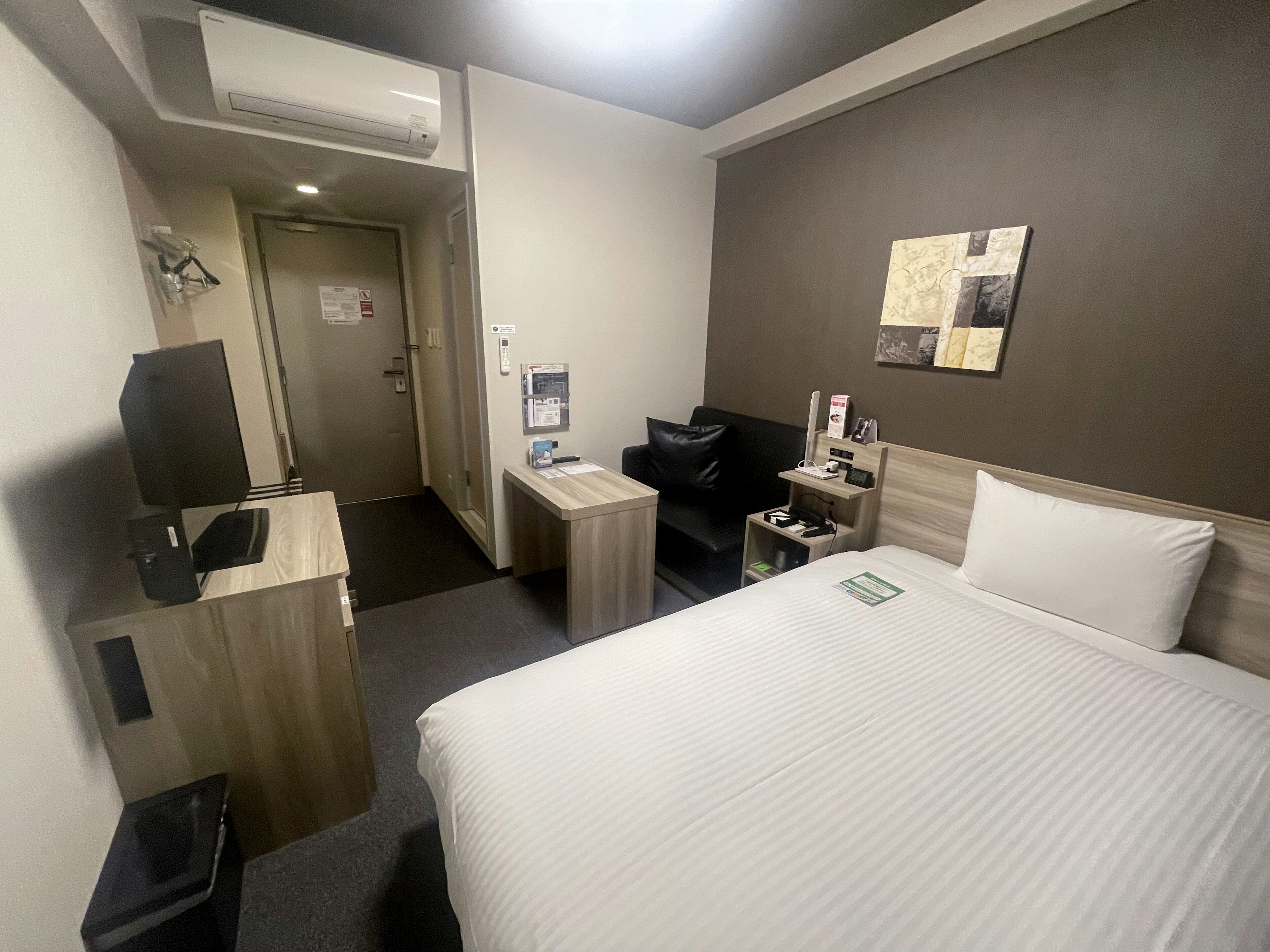 A hotel room with a bed, a desk, a chair, a TV, and a small table with a lamp.