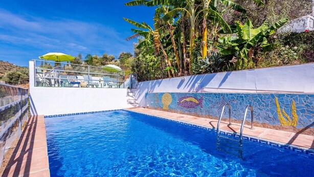 Une piscine avec un mur décoré de mosaïques, des chaises longues et un espace salon abrité.