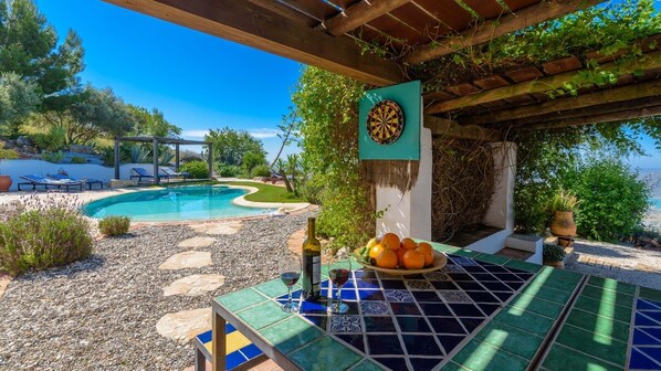 Zona junto a la piscina con dardos, una botella de vino y un bol de naranjas sobre una mesa de baldosas.