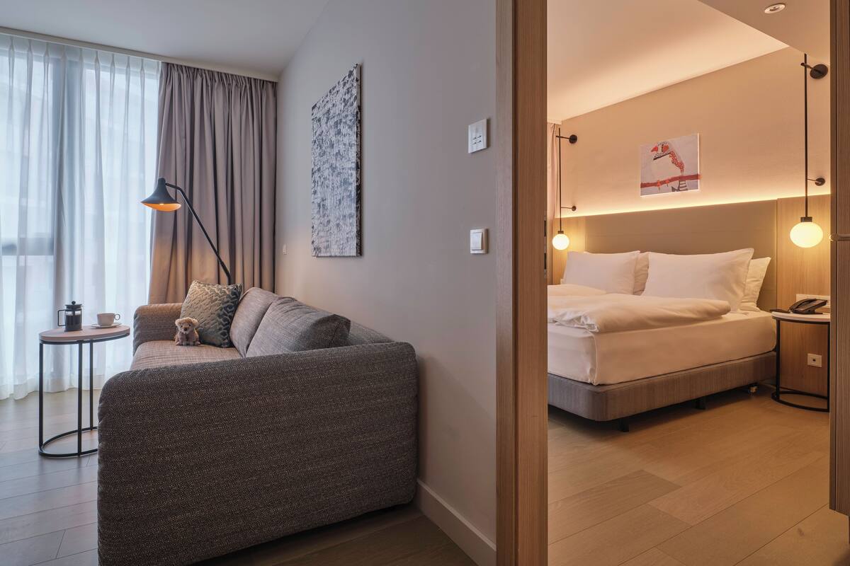 Una habitación de hotel moderna con sofá, una mesita con cafetera, cama con ropa de cama blanca y dos lámparas de noche.