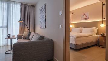 A modern hotel room with a sofa, a small table with a coffee pot, a bed with white bedding, and two bedside lamps.