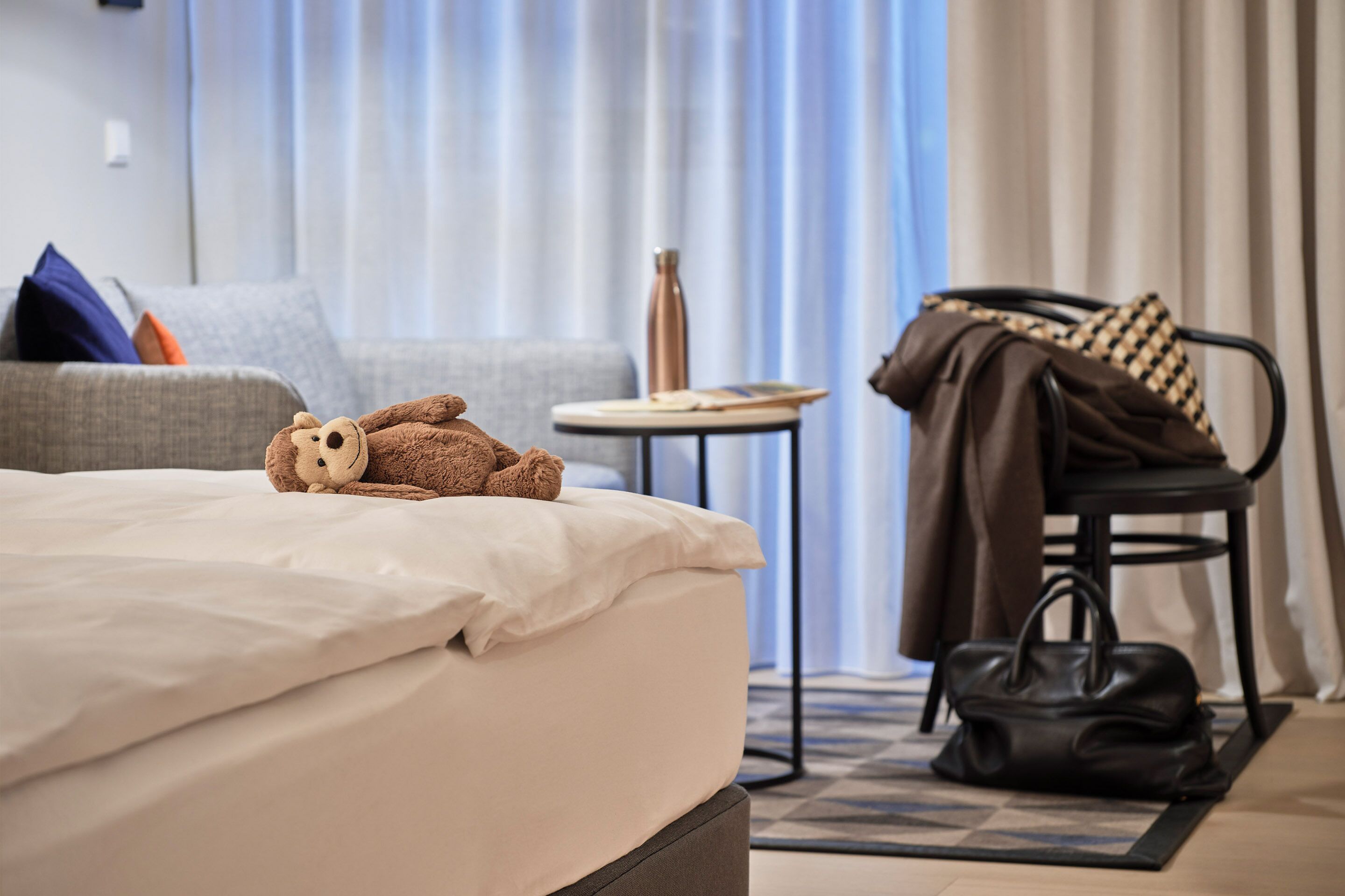 A neatly made bed with a teddy bear, a grey sofa with cushions, a small round table with a bottle, and a chair with a coat and bag.