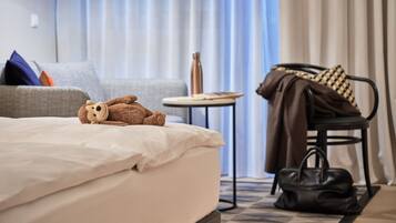 Una cama bien hecha con un osito de peluche, un sofá gris con cojines, una mesita redonda con una botella y una silla con abrigo y bolso.